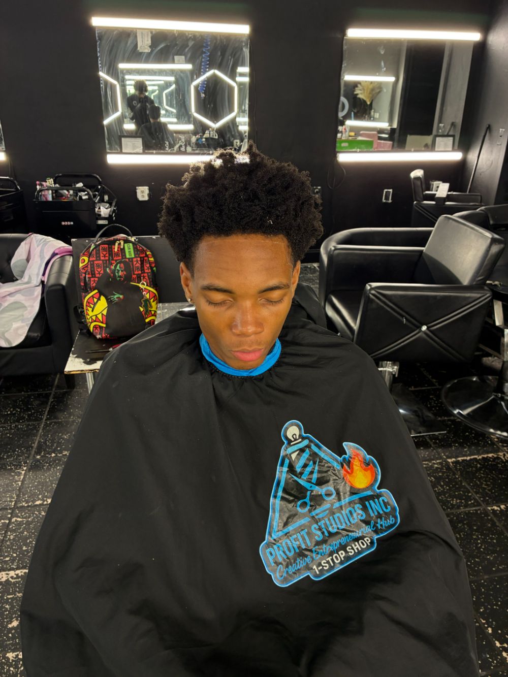 A young man in a Profit Studios cape sitting in the chair, with the empty chairs and mirrors behind him.