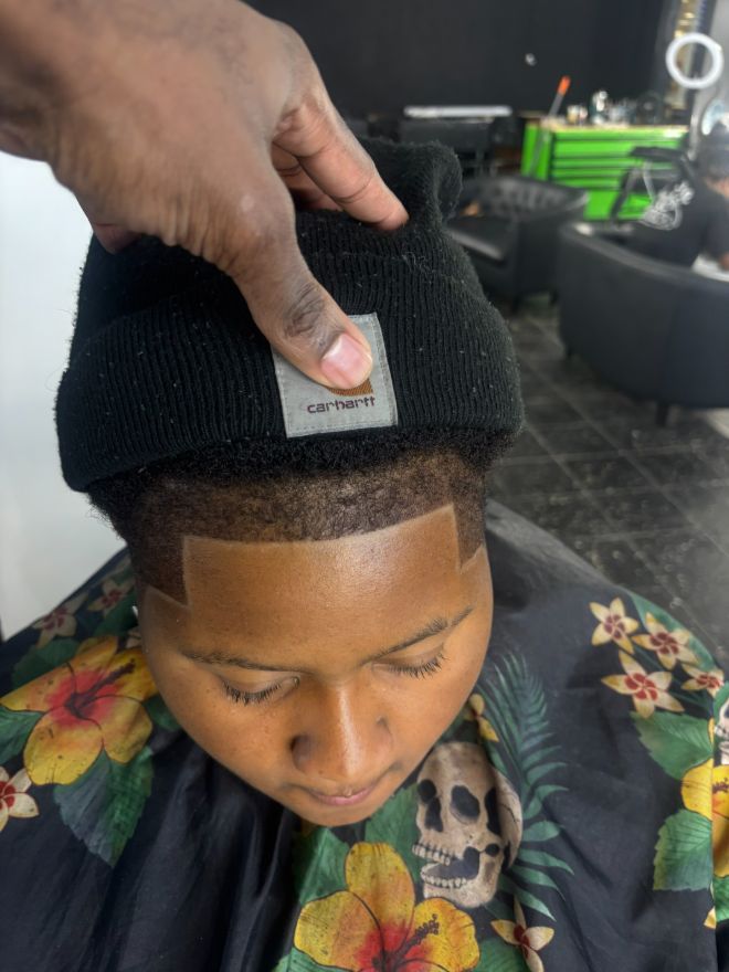 A hand lifting a beanie off a child's freshly cut hair while he sits in a floral cape.