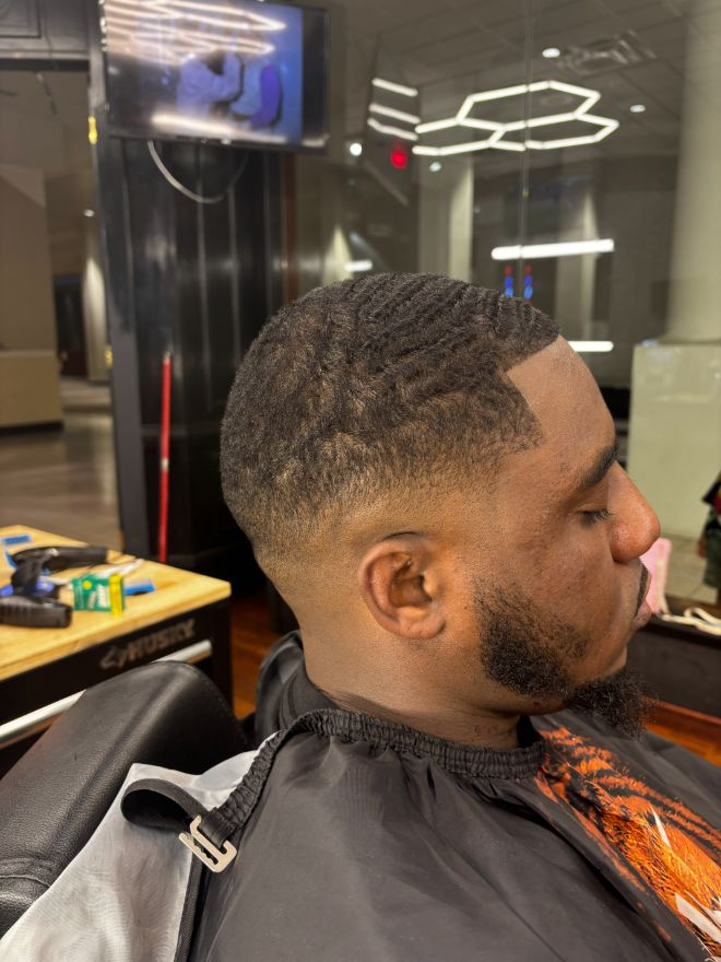 A man in a barber cape with brushed waves on top and a high fade, under the shop's hexagon lights.