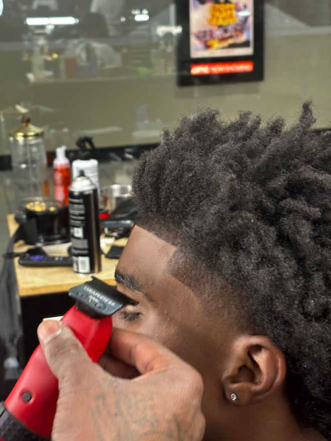 A close view of a barber's hand running red clippers along a client's hairline.